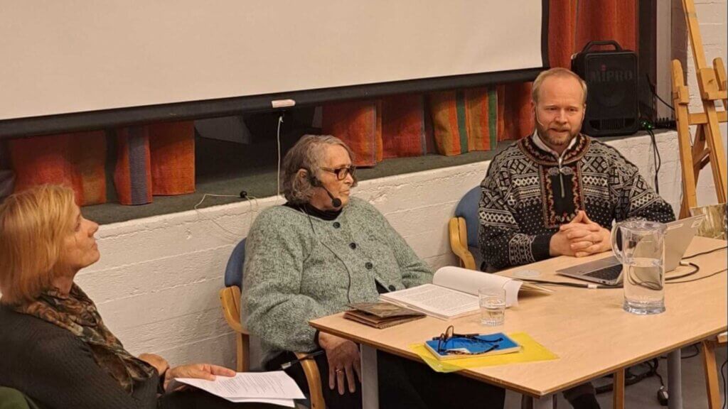 Tre personer deltar i en paneldiskusjon ved et bord med bøker og dokumenter. En mann bærer en tradisjonell genser.