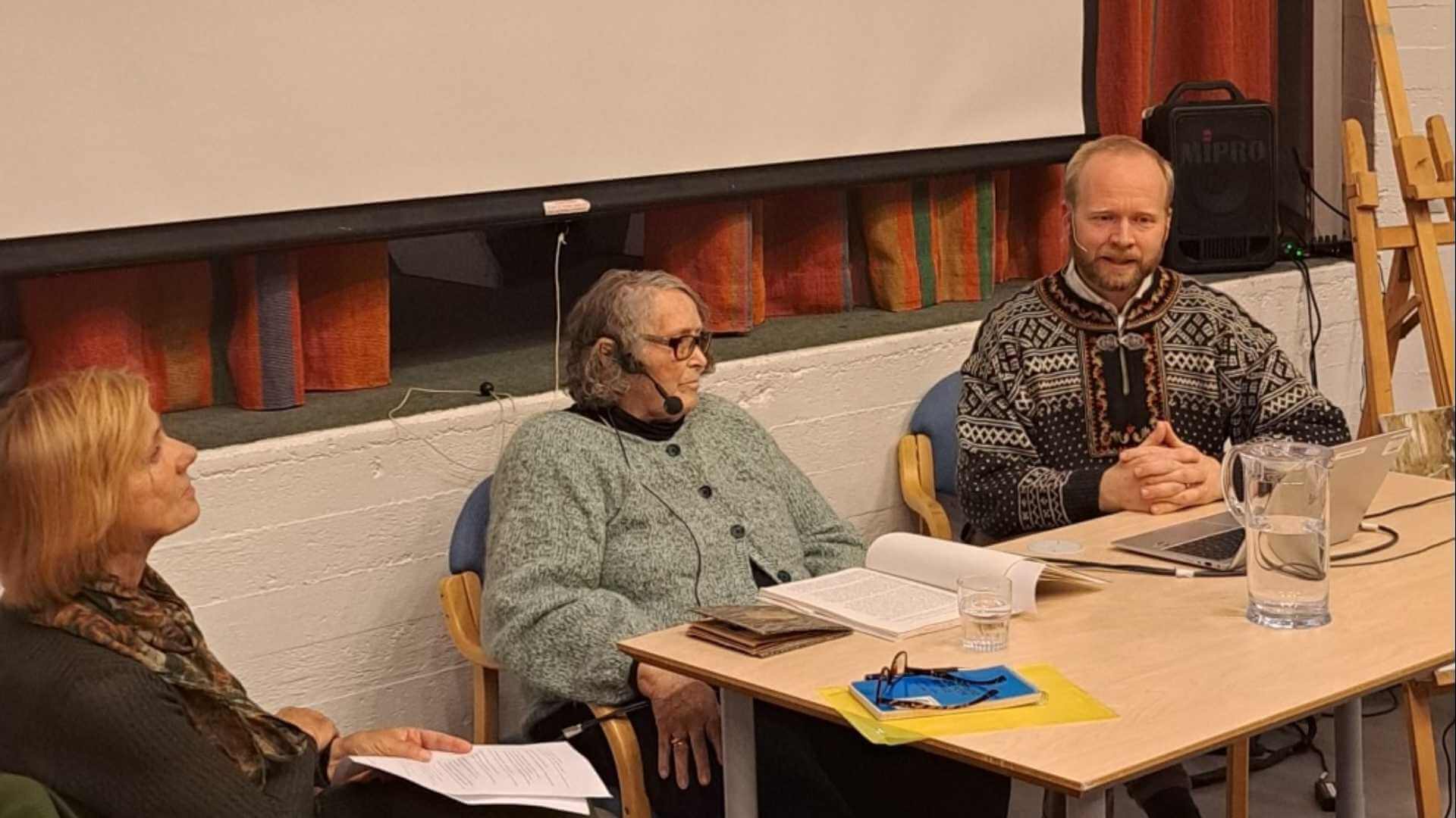 Tre personer deltar i en paneldiskusjon ved et bord med bøker og dokumenter. En mann bærer en tradisjonell genser.