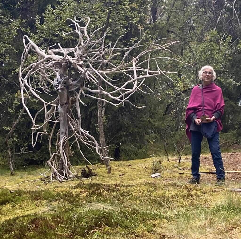 Eldre kvinne i rosa sjal står ved siden av et unikt, vridd tre i skogen.