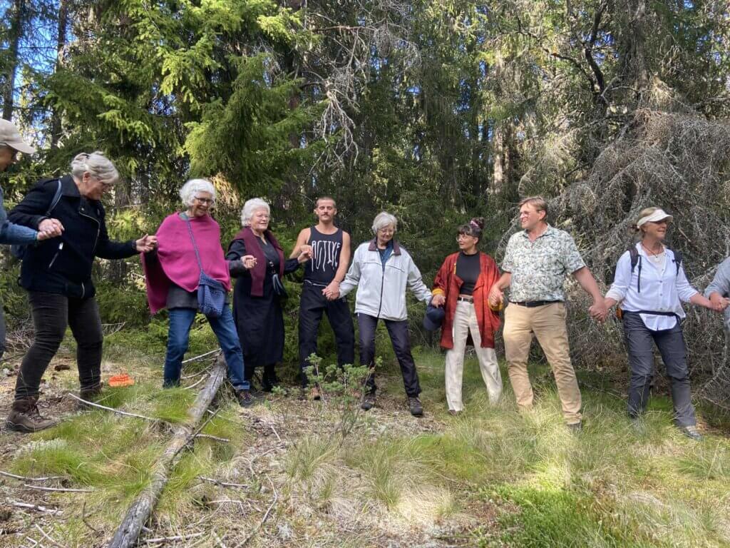 Ei gruppe menneske dansar ringdans i ei skog, omringa av grøn vegetasjon.