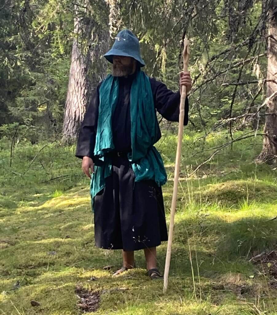 Mann med grønn kappe og stav i skogen på solrik dag.