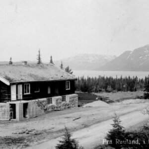 Gammelt ungdomshus i fjellandskap, med trær og fjord i bakgrunnen i Retland, Norge. Svart-hvitt bilde.