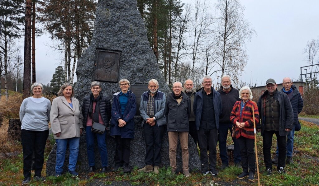 Gruppe mennesker foran en minnesmerke i skogsområde, omgitt av trær og høstlandskap.