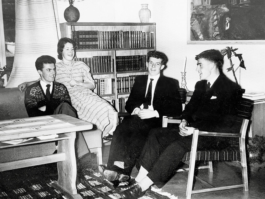 Fire unge mennesker i formelle antrekk sitter og prater i en stue med bøker i bakgrunnen.