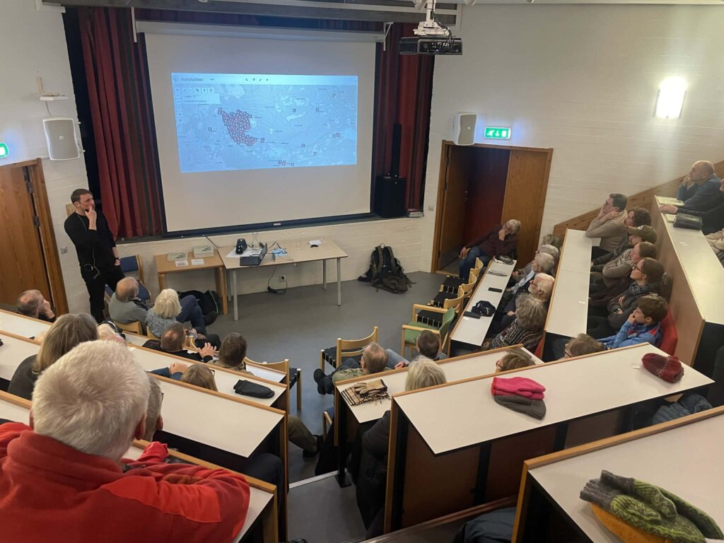 Folk samles i et klasserom for å høre på en presentasjon med kartvisning. Engasjert deltakelse.