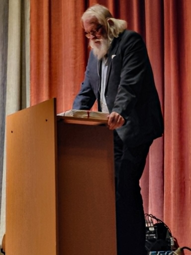 En eldre mann med skjegg står ved en talerstol, leser fra en bok foran et publikum med gardiner i bakgrunnen.