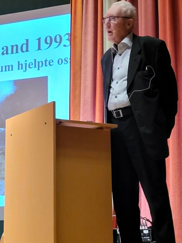 Elderly speaker at a podium discussing an event from 1993, with a presentation screen in the background.