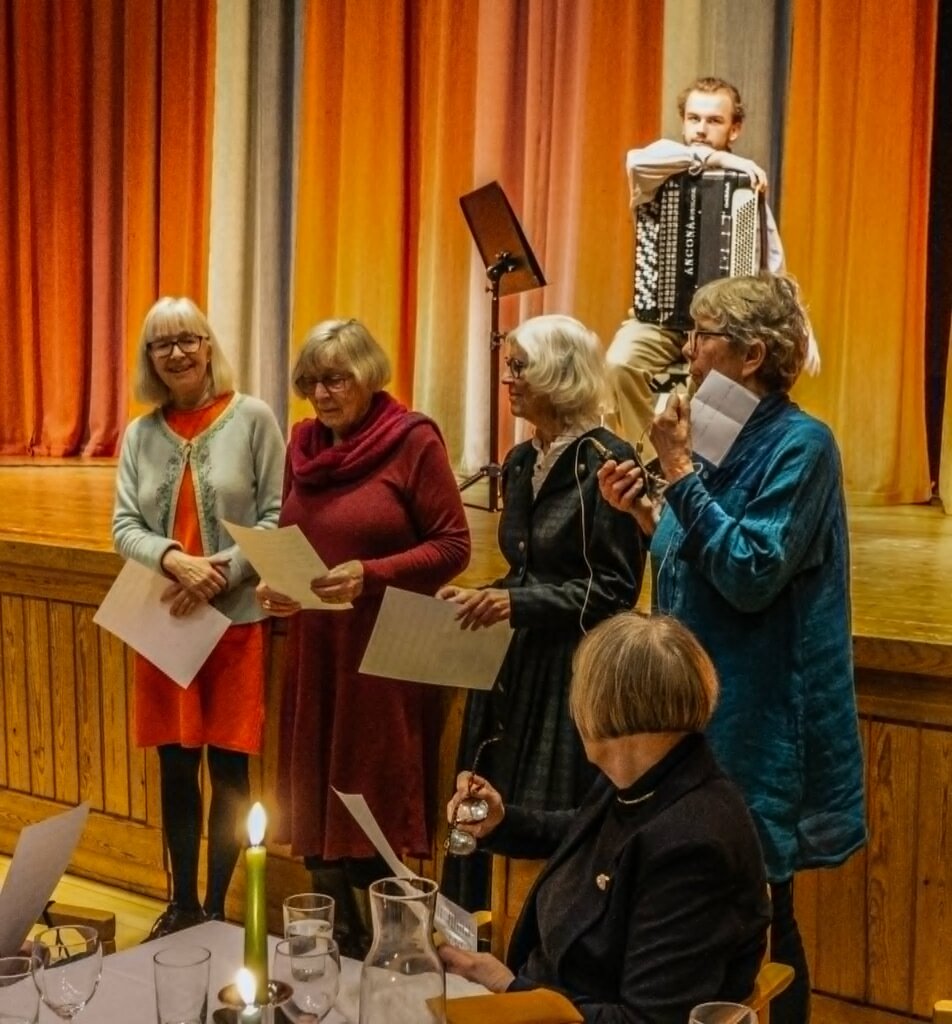 Fem kvinner står på en scene med manuskript mens en musiker spiller trekkspill i bakgrunnen.