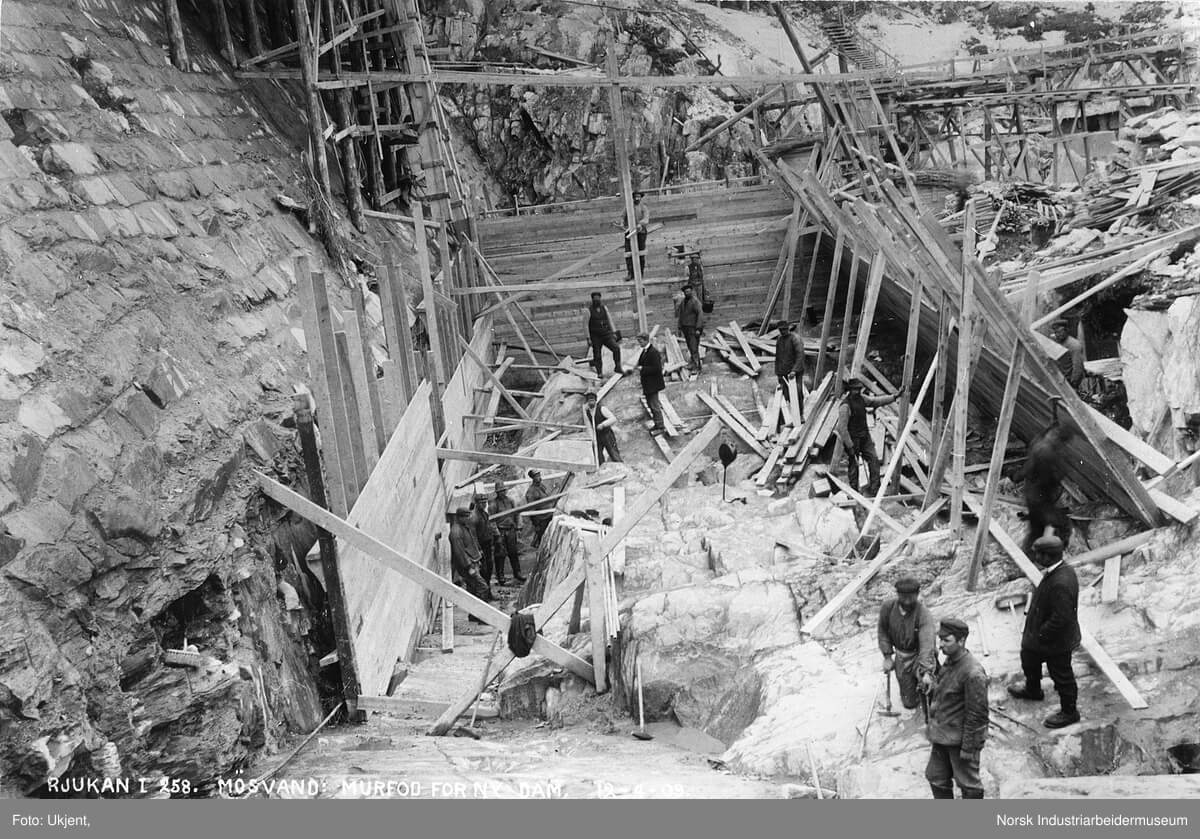 Historisk bilde av bygging av Murfossdammen i Rjukan, med arbeidere og treverk i konstruksjonen.