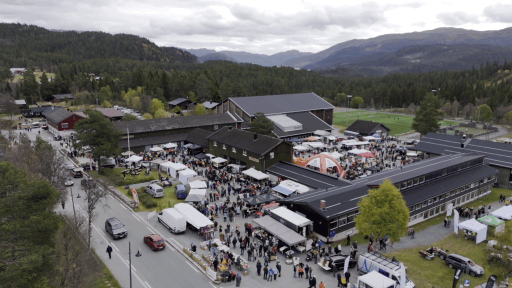 Folkemengde på en utendørs festival i naturskjønne omgivelser med fjell og skoger i bakgrunnen.