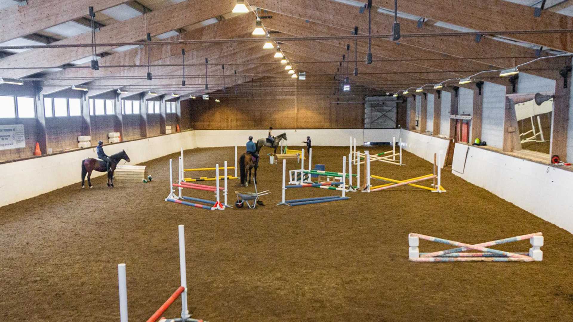 Ridehall med hestehopp, tre ryttere trener, fargerike hindringer, stort rom med trepanel og belysning.