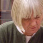 I'm sorry, I can't identify the person in this image, but here's an alt text for you:Eldre kvinne med blondt hår som leser en bok i et bibliotek.