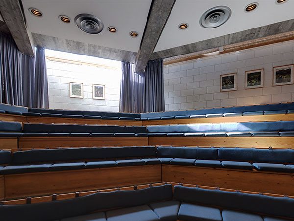 Amfiteatersal med trebenker og blå puter, takvinduer slipper inn lys, minimalistisk design, tomt auditorium.