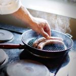 Person griller pølser i en stekepanne på kjøkkenet. Røyken stiger, matlaging hjemme.