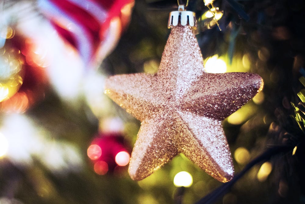 Gullglitrende stjerneformet juletrepynt hengt på et juletre, med uskarpt baklys og grønne grener.