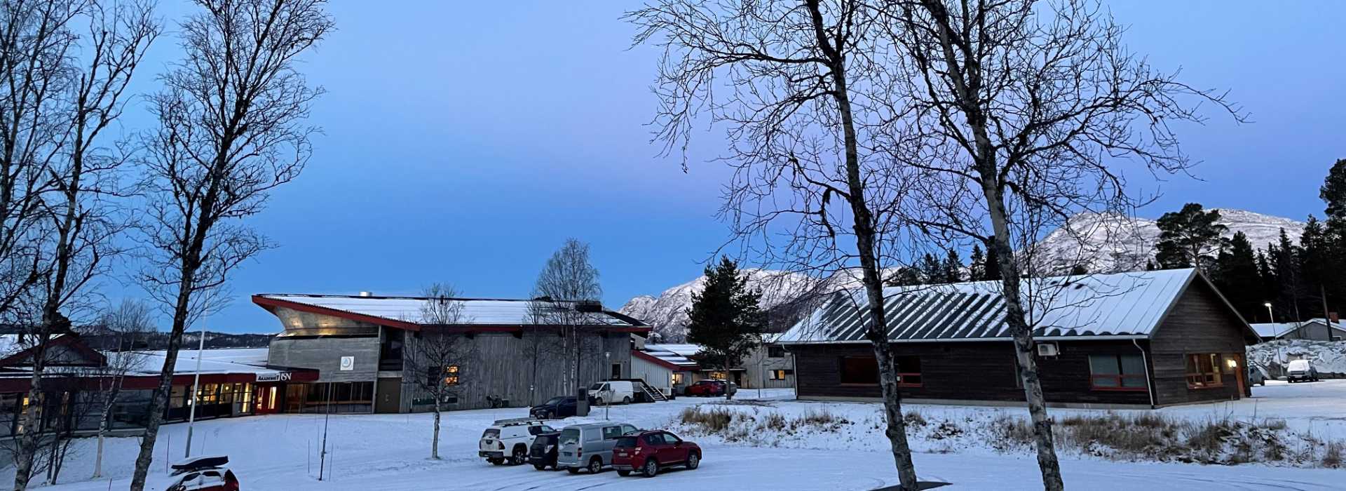 Vinterlandskap med snødekte bygninger, trær og fjell i bakgrunnen under den blå himmelen.
