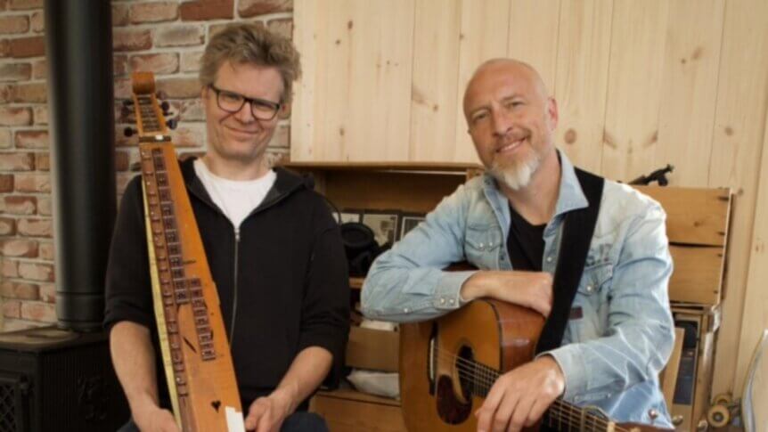 To menn med musikkinstrumenter, en har en gitar og den andre en langeleik. Brick vegg og trepanel i bakgrunnen.