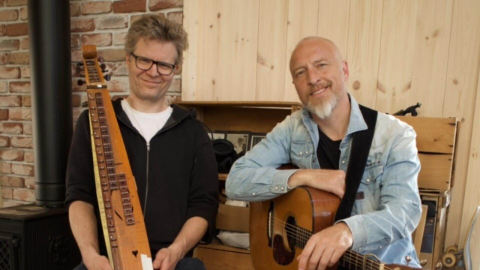 To menn med musikkinstrumenter, en har en gitar og den andre en langeleik. Brick vegg og trepanel i bakgrunnen.