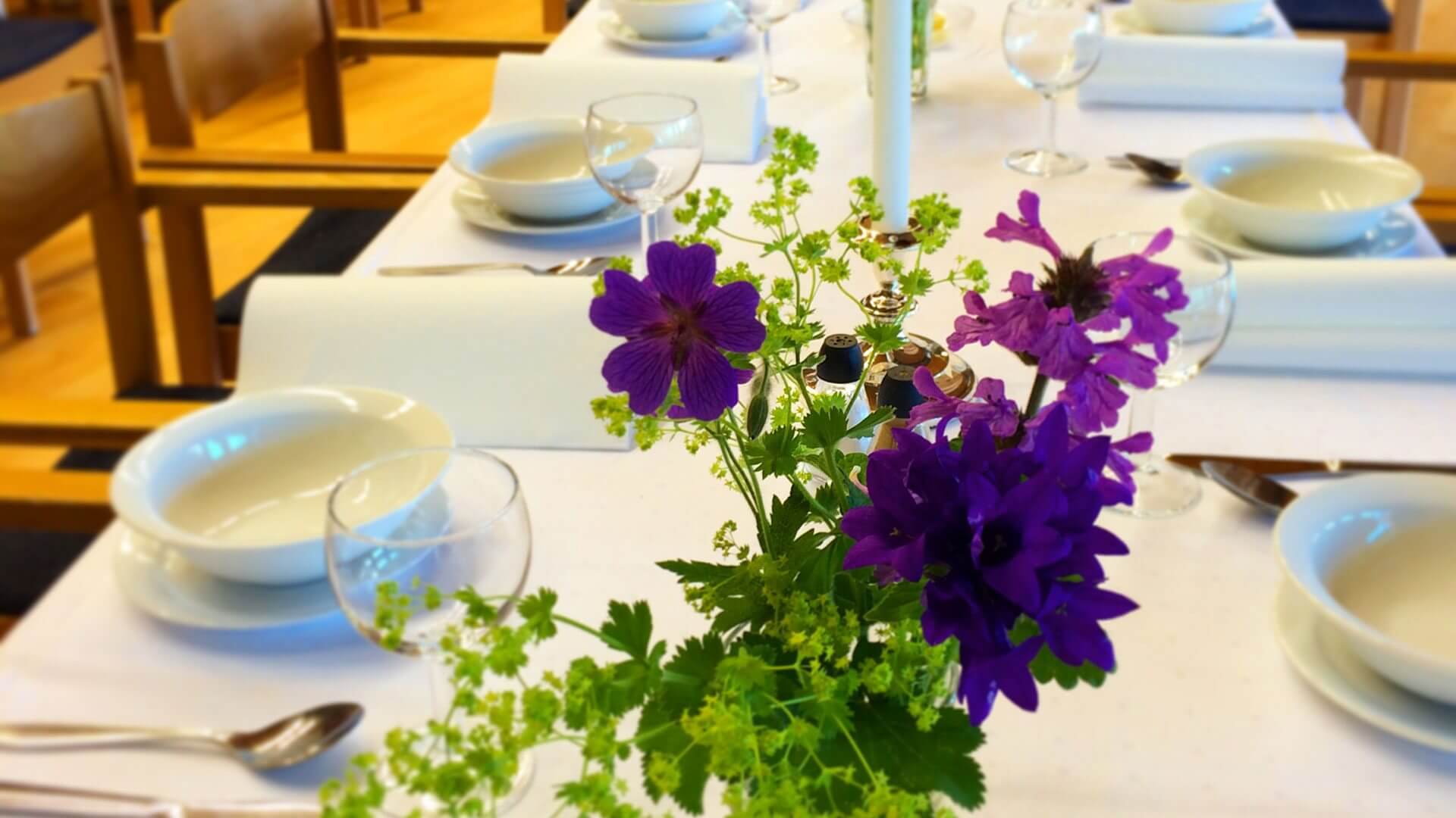 Borddekorasjon med lilla blomster og dekketøy på hvit duk, klar til selskap i en elegant spisesal.