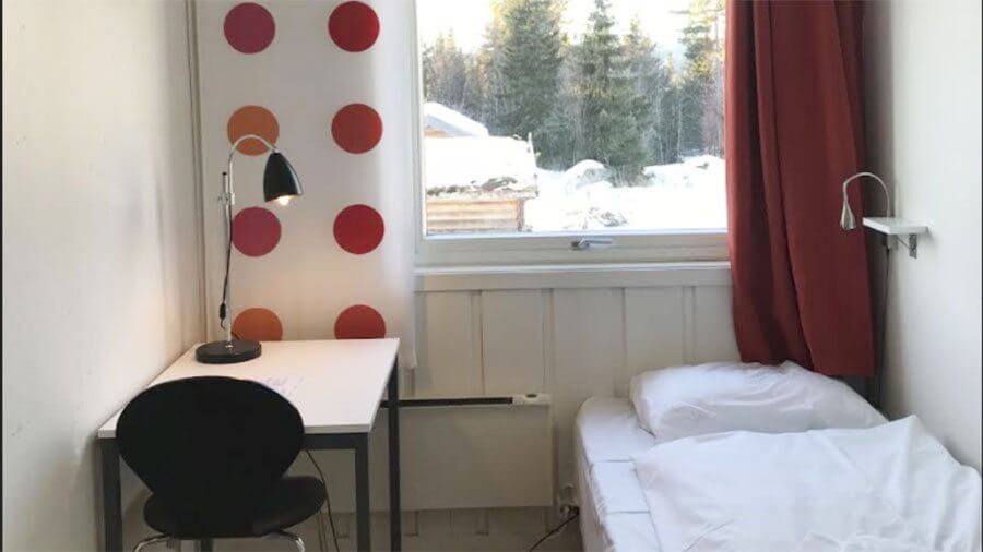 Hyggelig rom med skrivebord, stol og seng ved siden av stort vindu. Utsikt til snødekket landskap og skog.