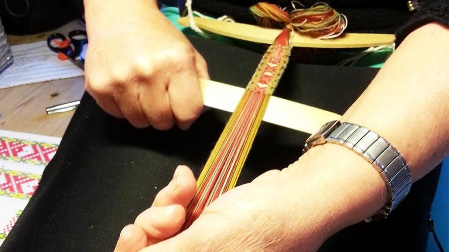 Person vever tradisjonelt bånd med grindvev, nærbilde av hendene i arbeid med fargerike tråder.