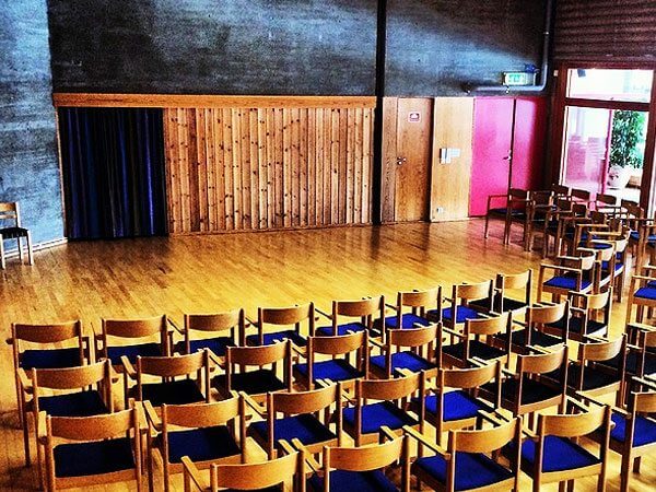 Rader med trestoler i en tom sal med tregulv og mørke vegger, klare for et arrangement.