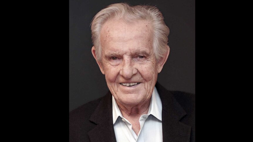 Eldre mann med grått hår og smilende ansikt, iført svart jakke og lys skjorte, foran mørk bakgrunn.