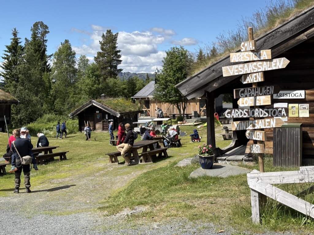 Besøkende nyter en solfylt dag i naturen med bygninger og informasjonsskilt i bakgrunnen.