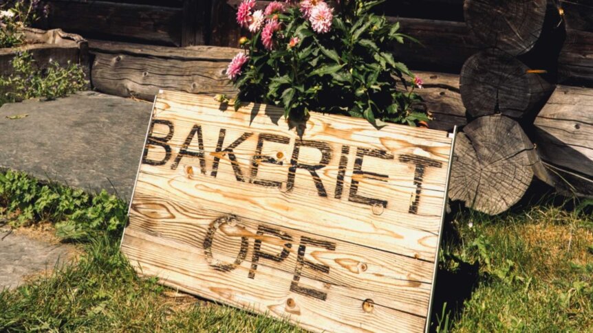 Skilt med teksten Bakeriet Åpent og blomster, plassert ved en trevegg i en grønn hage.