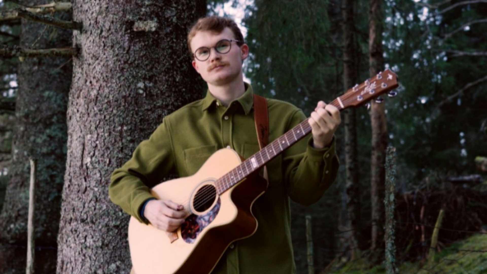 Ung mann med briller spiller gitar i skogen, kledd i grønn skjorte mot en bakgrunn av trær.