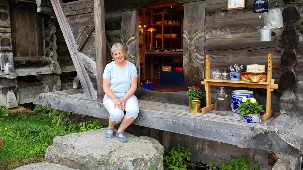 Kvinne sitter på en stein ved et trehus, med blomster og servise i bakgrunnen. Norsk kulturarv og håndverk.
