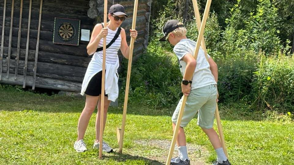 To personer som prøver stokker for å gå på ski i en solfylt hage, med en hytte i bakgrunnen.