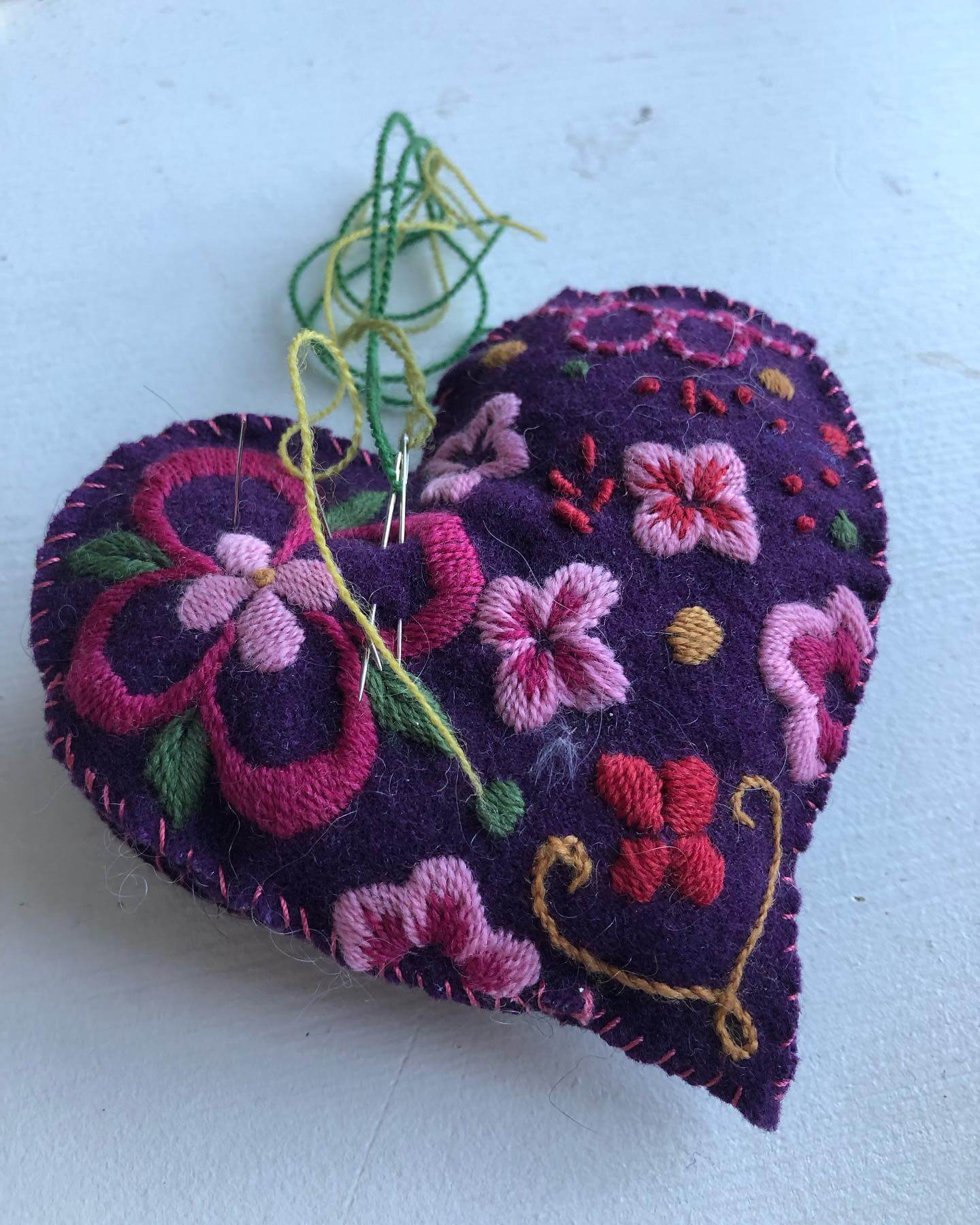 Hjerteformet nålepute med lilla bakgrunn og fargerike blomsterbroderier, perfekt for syglede.