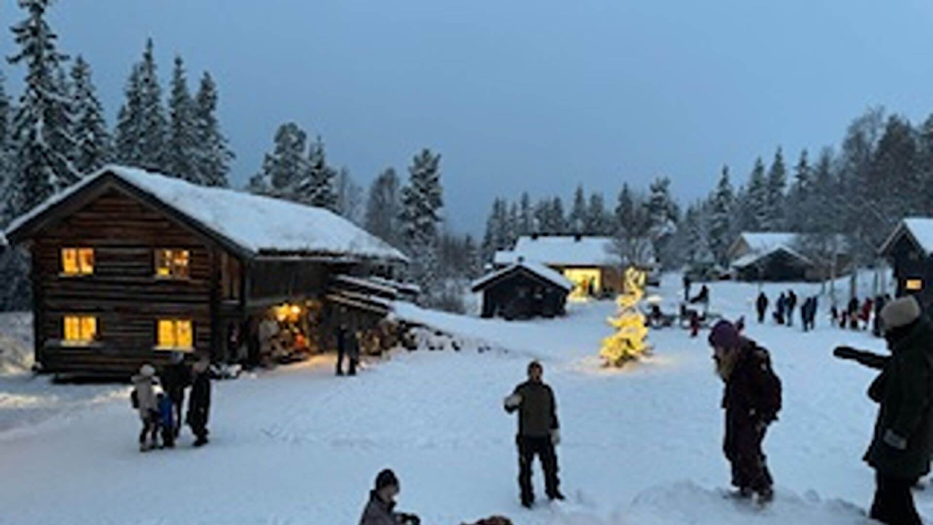 Familier nyter snødekt landskap ved hytter, med juletre og folk i vinterklær. Koselig atmosfære i nydelig vinterlys.