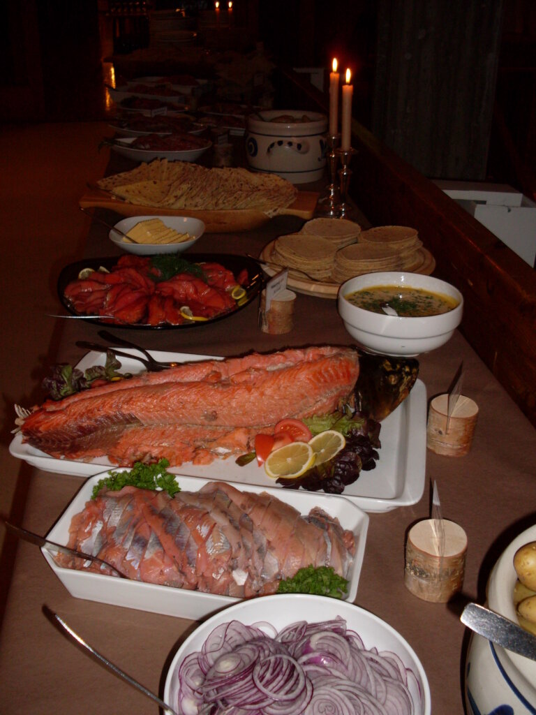 Alt tekst: Et vakkert dekket bord med ulike fiskeretter, brød, løk, og grønnsaker i en lun atmosfære med stearinlys.