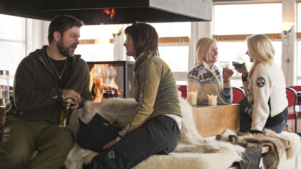 Gruppe som koser seg foran peisen, drikker og prater i varm og hyggelig atmosfære i vinterhyttemiljø.