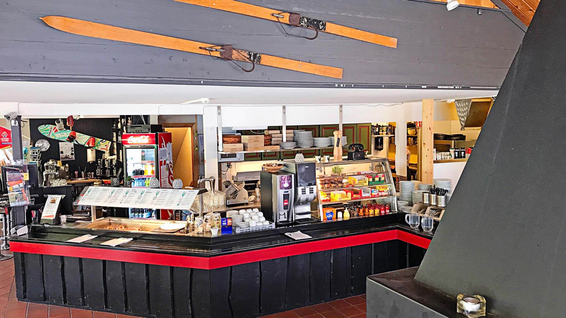 Kafeteria med servering av mat og drikke under gamle ski, viser rustikk interiør og menyer på disken.