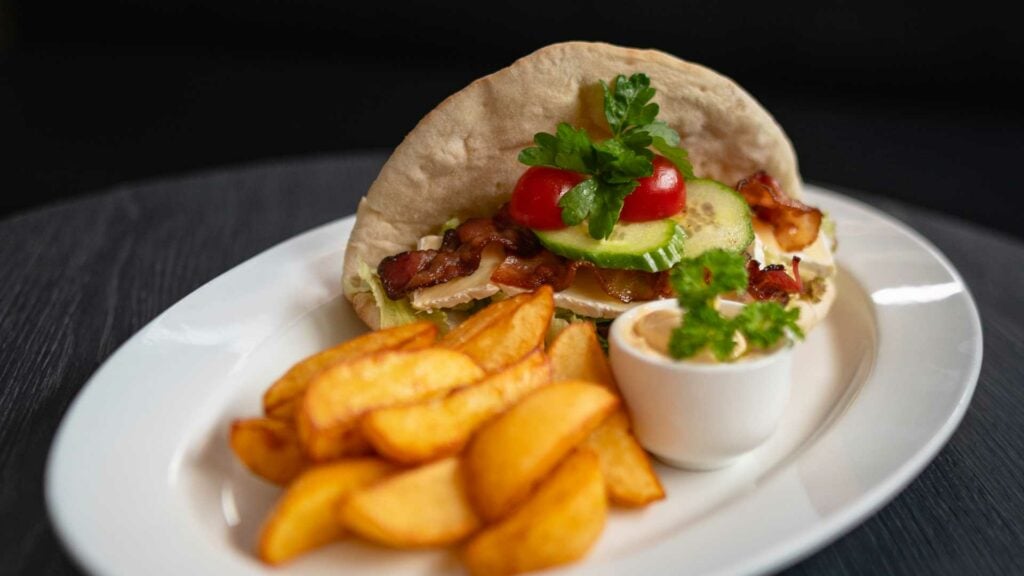 Pitaburger med bacon, agurk og tomat, servert med potetbåter og aioli på en hvit tallerken.