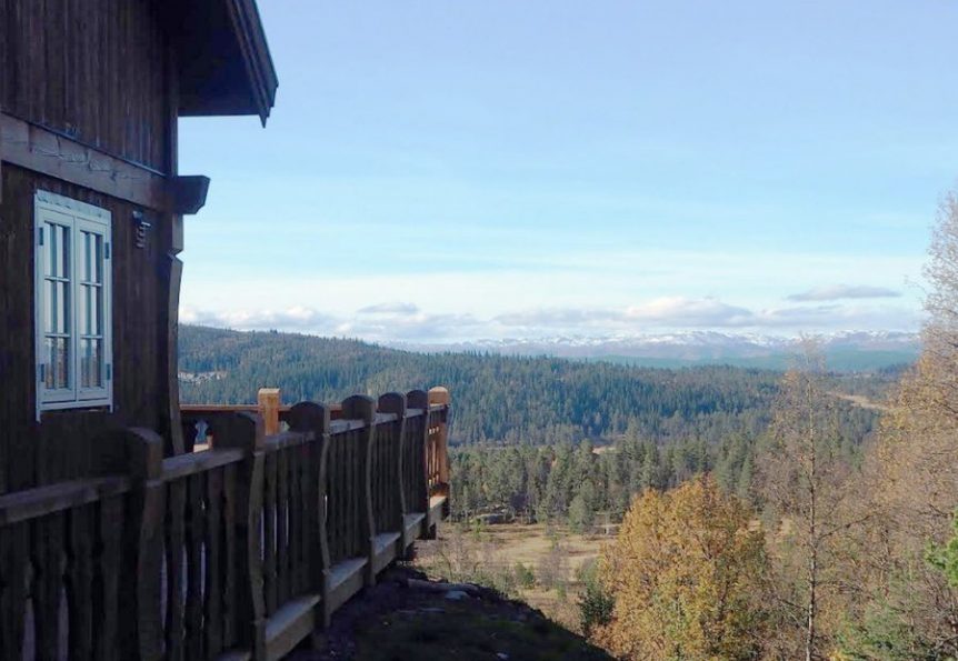 Tre hytte med utsikt over skogkledd dal og fjell i bakgrunnen under en blå himmel.