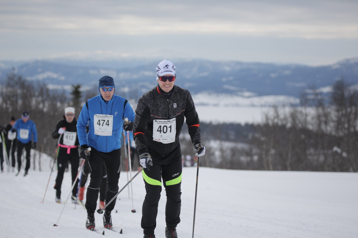 Flere langrennsløpere konkurrerer i et vinterlandskap, bakkene dekket av snø foran fjell i horisonten.