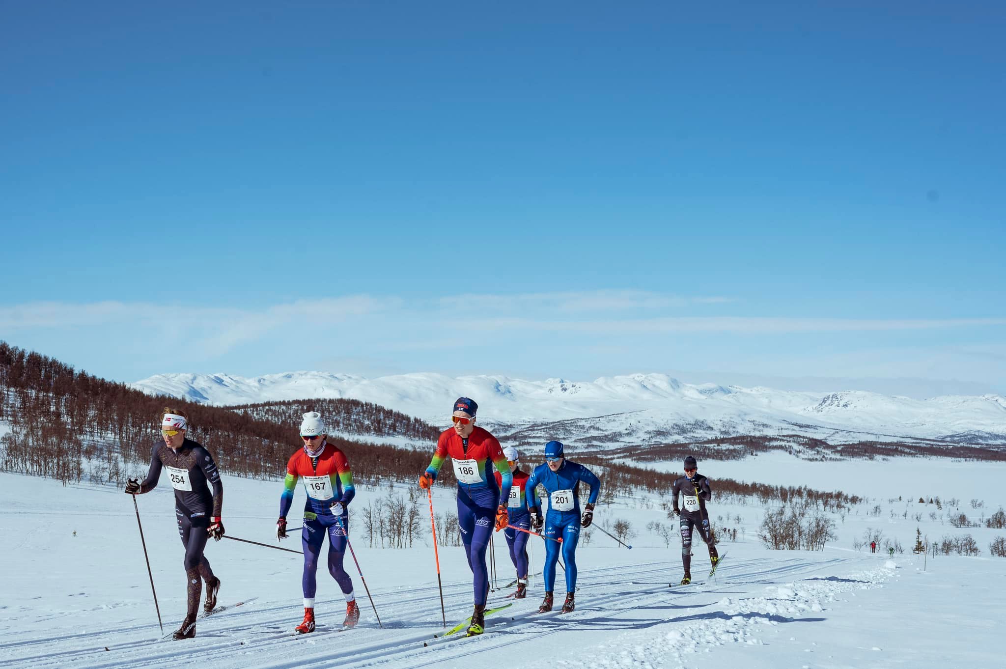 Langrennsløpere i konkurranse på snødekte fjell, klar blå himmel og majestetisk bakgrunn i Norge.
