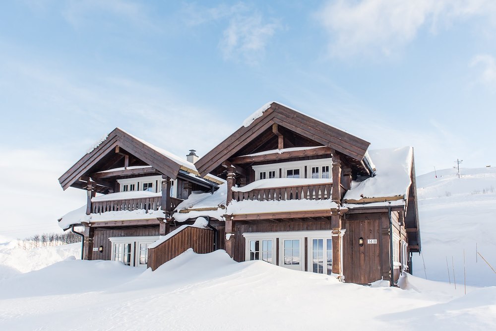 Trebygget hytte dekket av snø i vinterlandskap under blå himmel.