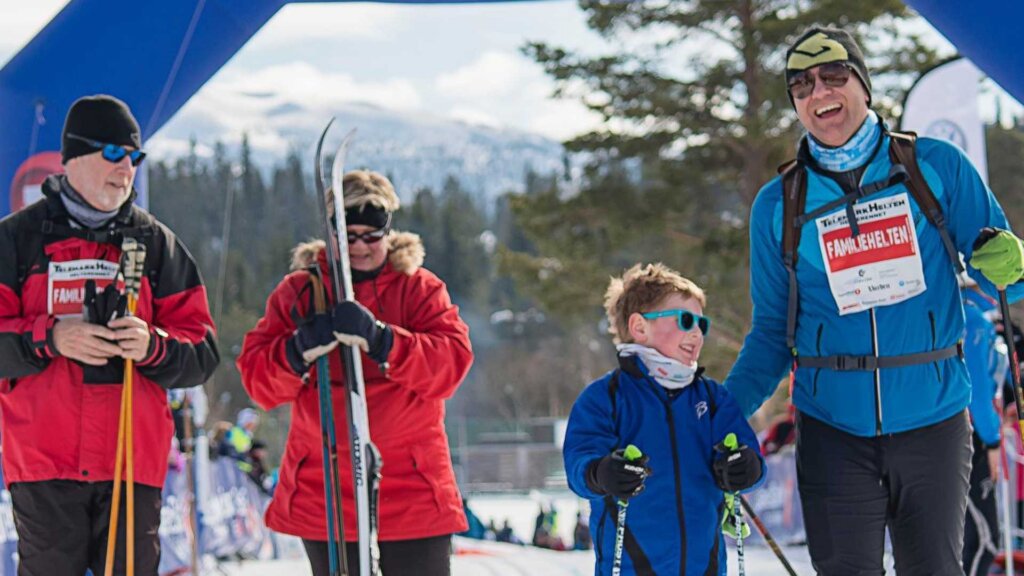 Familie på skitur, smilende i snødekte omgivelser, løper under et blått målseil i fjellet.