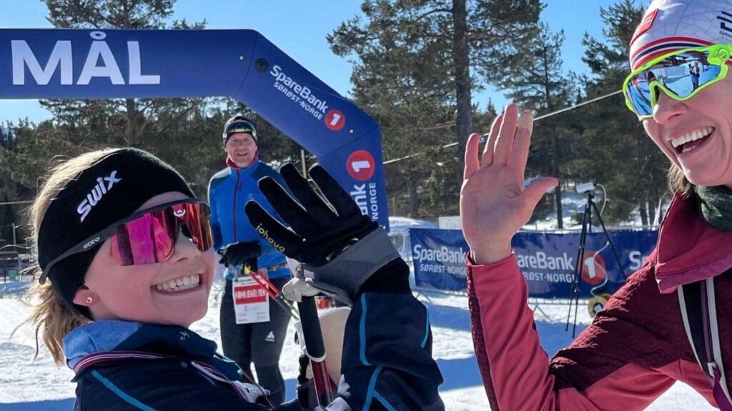 To glade skiløpere gir hverandre en high-five ved mållinjen under et skirenn på en solfylt dag.