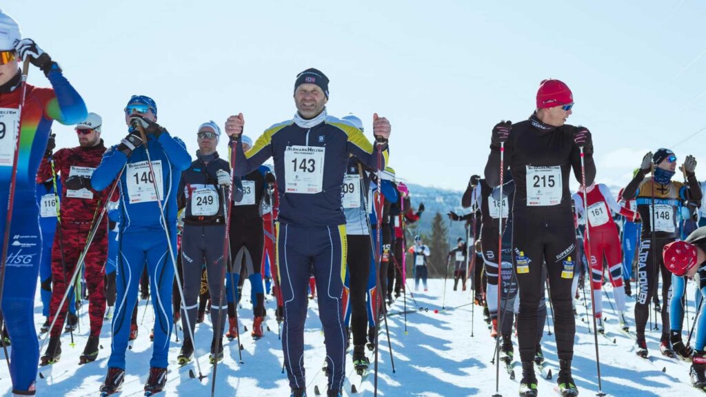 Langrennsløpere klare til start i solfylt vinterlandskap under skirenn, iført racingdrakter og startnummer.