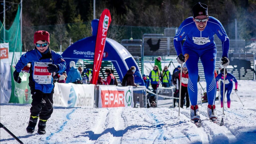 To personer på ski i en konkurranse under blå himmel, omgitt av bannere og tilskuere.
