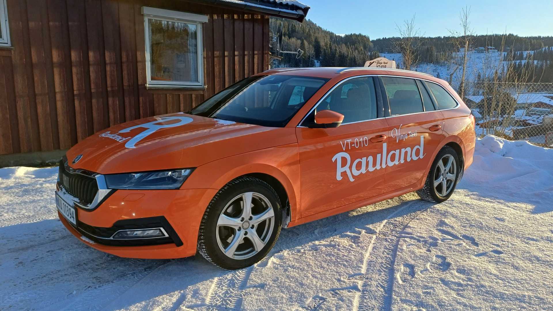 Oransje taxi med Rauland parkert i snødekt område foran en trebygning.