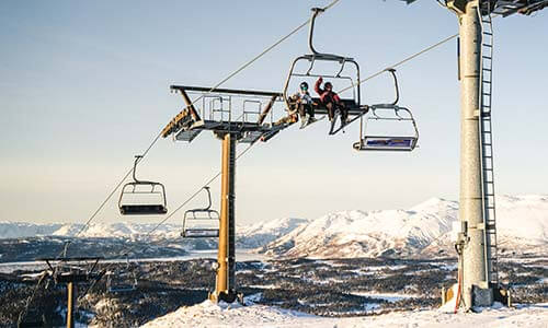 Personer på stolheis i snødekt fjellandskap med klar himmel i bakgrunnen. Vinter og skianlegg i Norge.