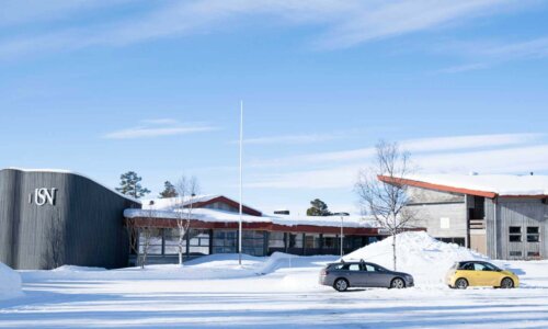 Moderne universitetsbygning dekket av snø, med flere parkerte biler foran, på en solrik vinterdag.