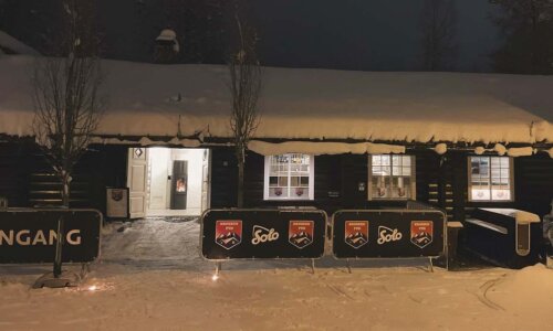 Koselig pub med snødekket tak og inngang opplyst om natten i Norge. Skilt viser vei til inngangen.